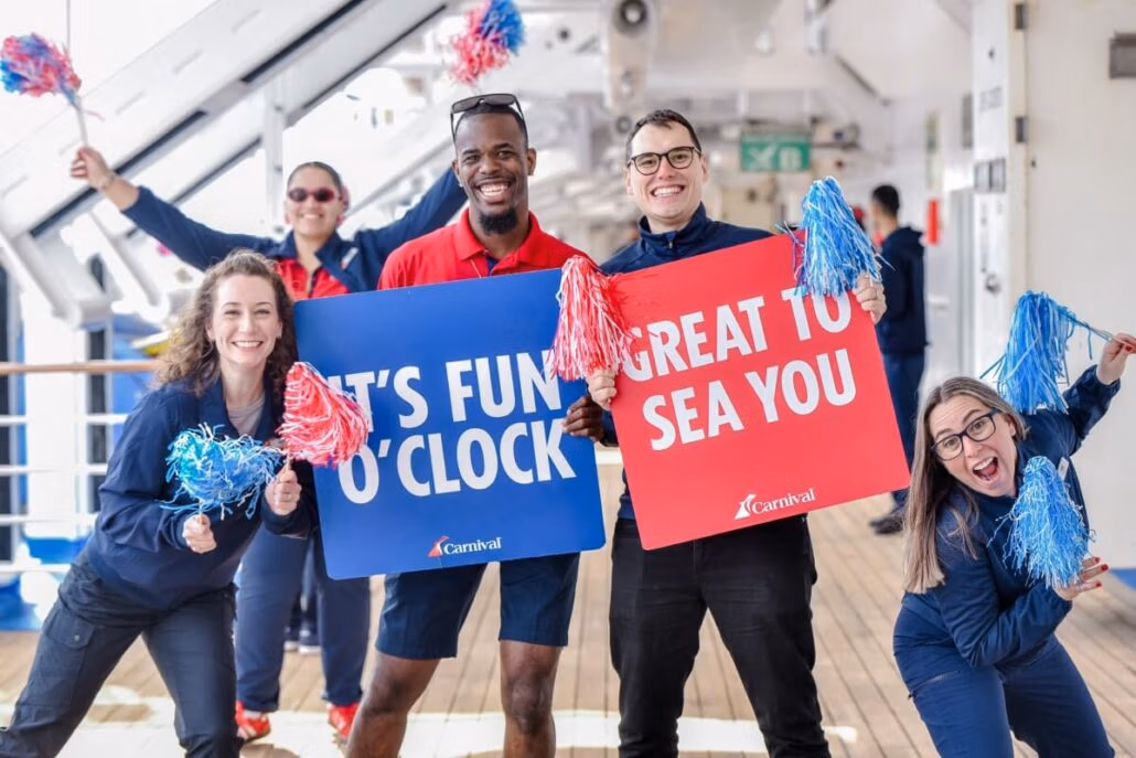 Five people in uniforms hold pom-poms and two signs, "IT'S FUN O'CLOCK" and "GREAT TO SEA YOU," on the Carnival Spirit's deck. Smiling warmly, they seem to be part of a welcoming crew celebrating the ship's return to service after dry dock.