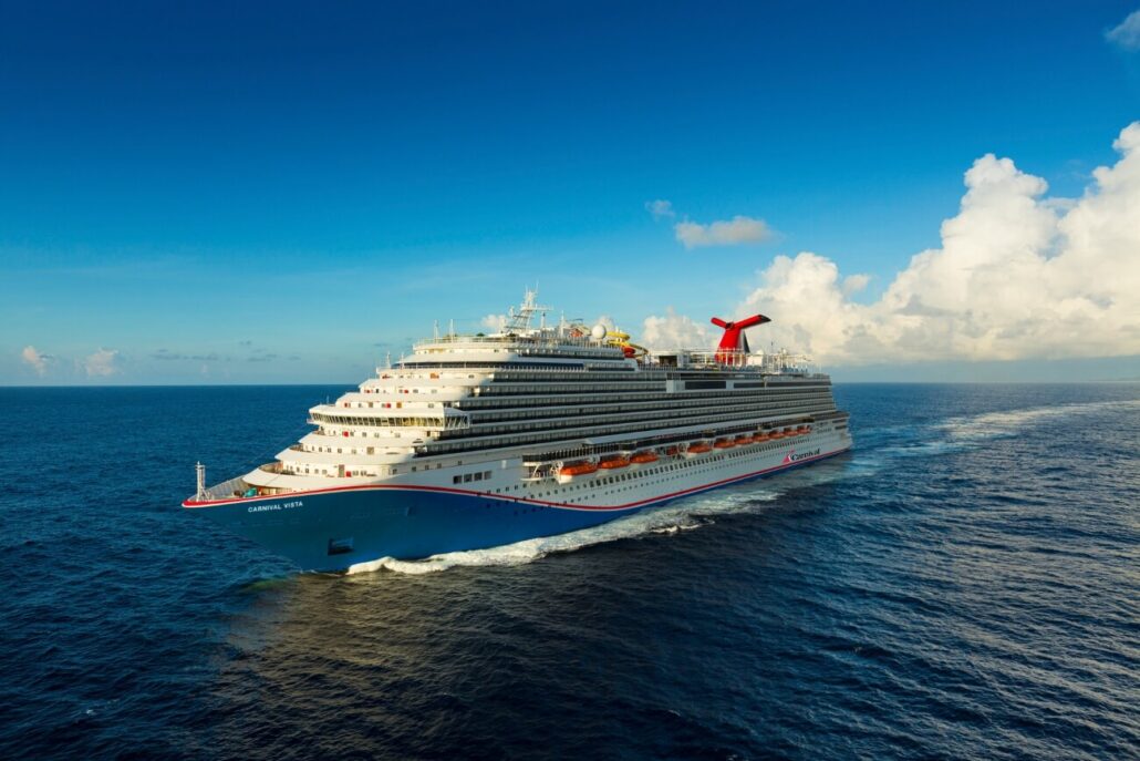 A family-friendly Caribbean cruise ship sails on a vast ocean under a clear blue sky, with white clouds in the distance. This impressive vessel boasts multiple decks and a distinctive red and blue funnel, while gentle waves trail behind as it journeys forward.