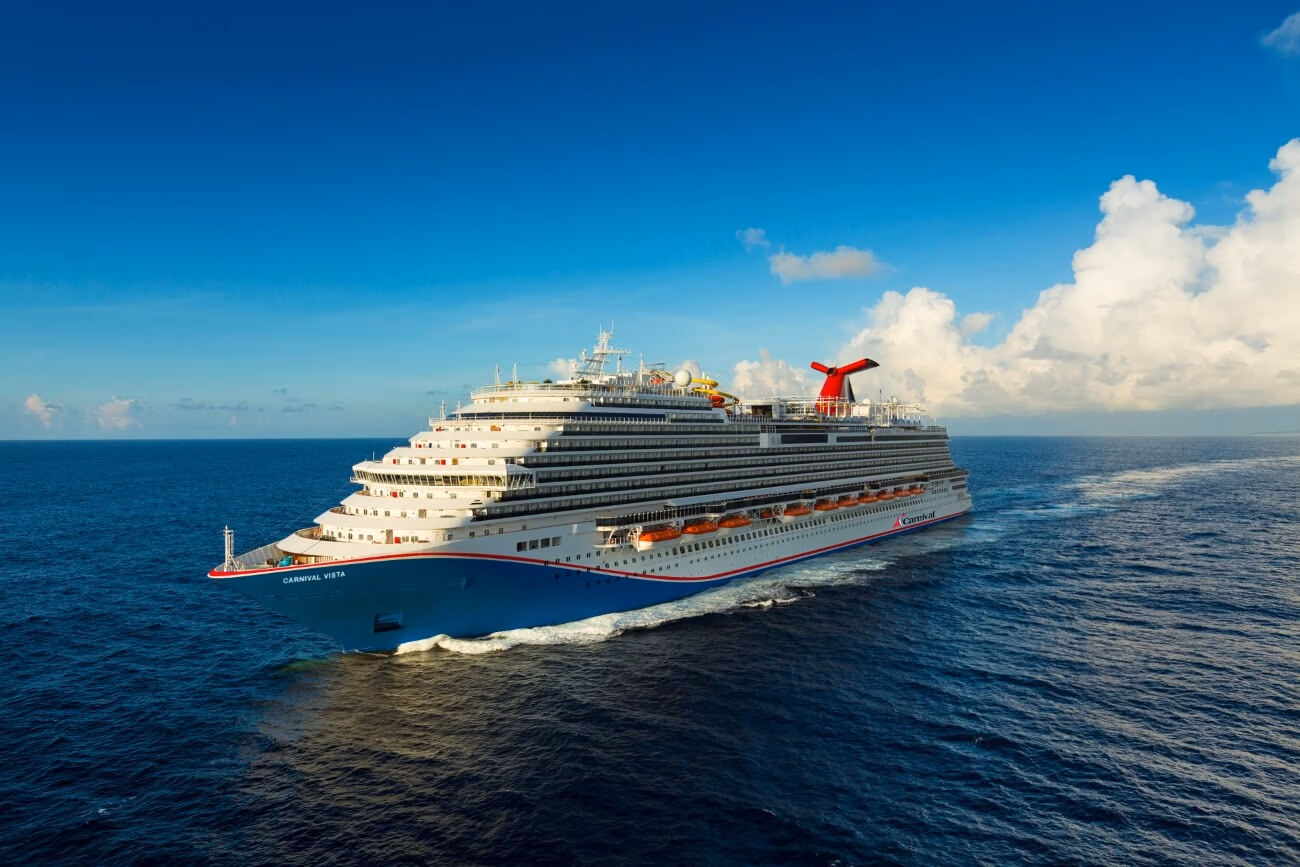 A family-friendly Caribbean cruise ship sails on a vast ocean under a clear blue sky, with white clouds in the distance. This impressive vessel boasts multiple decks and a distinctive red and blue funnel, while gentle waves trail behind as it journeys forward.