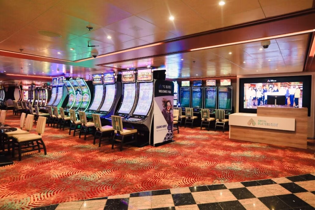 A casino interior featuring rows of slot machines with colorful screens on a patterned red carpet evokes a vibrant Carnival Spirit atmosphere. On the right, a digital display shows an image beside a counter with a service sign. The lively space is illuminated by ceiling lights.