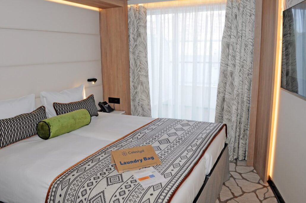 The hotel room offers premium accommodations, featuring a double bed with white and patterned bedding, multiple pillows including a green cylindrical one, and a wooden headboard. Enjoy a wall-mounted TV, curtains partially veiling the window, and discover Celestyal Discovery-level service.
