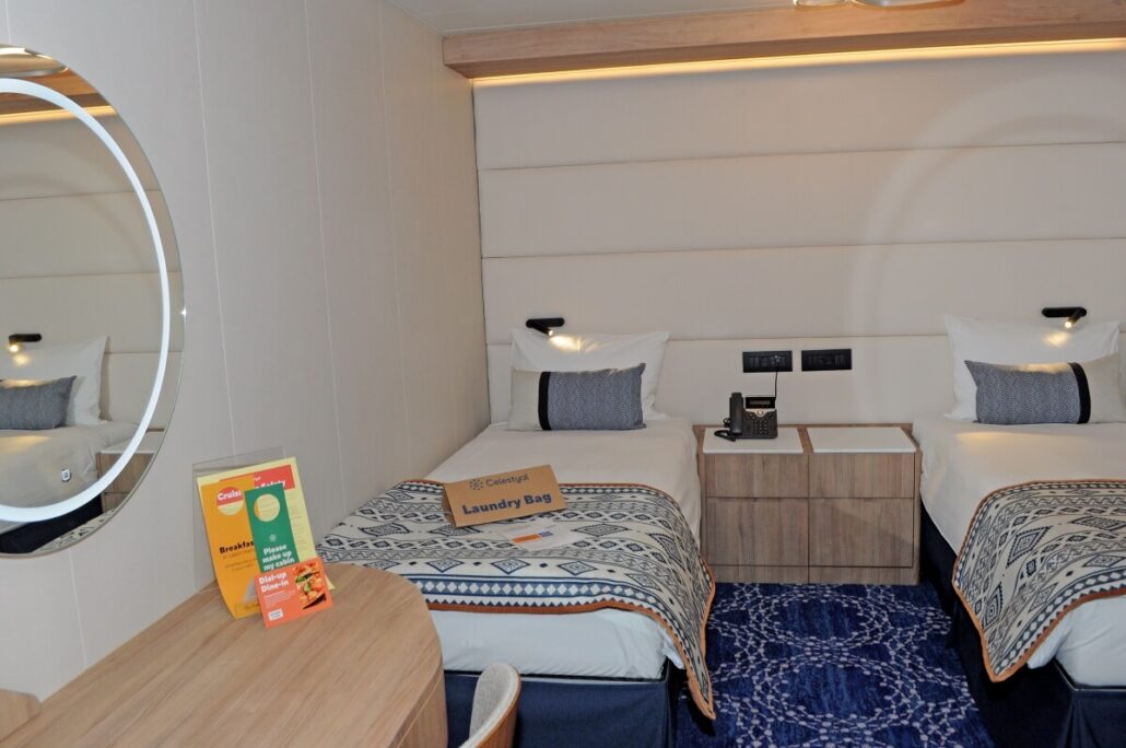 The Celestyal Discovery welcomes you with premium accommodations featuring two single beds adorned with patterned blankets and blue cushions. A wooden table, round mirror, brochures, laundry bag, and bedside telephone all enhance your stay.
