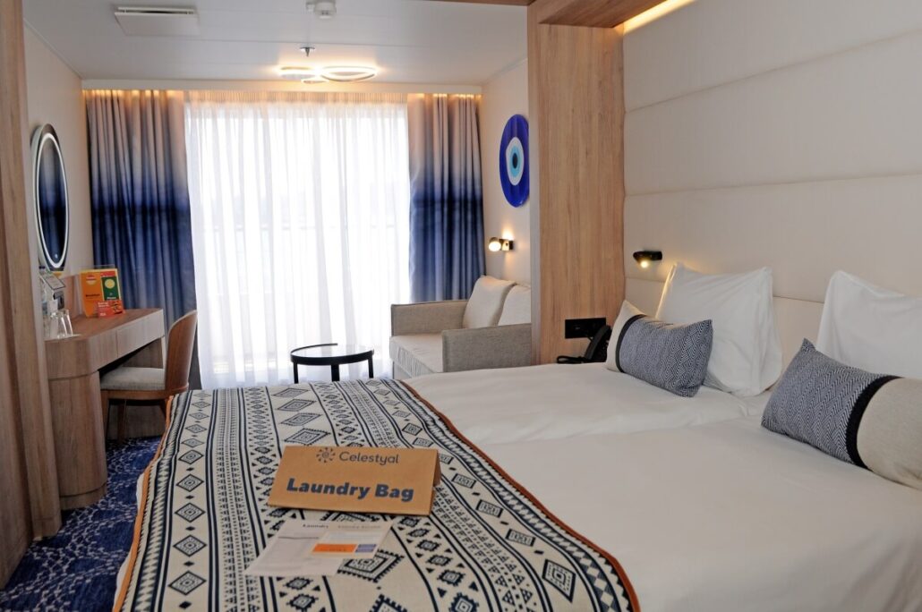 The hotel room offers premium accommodations with two neatly made twin beds adorned with patterned throws and pillows. To the left, a desk boasts a chair and lamp beneath a round mirror. Large windows invite natural light, while a "Laundry Bag" rests on one bed, ready for Celestyal Discovery's return to service.