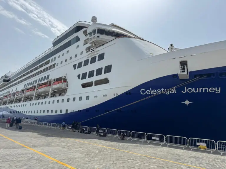 The majestic Celestyal Journey, a large white cruise ship with a striking blue hull, is docked at the port for Embarkation Day. Lifeboats line its side as people gather near security barricades on the pavement. The sky over Doha remains clear and bright.