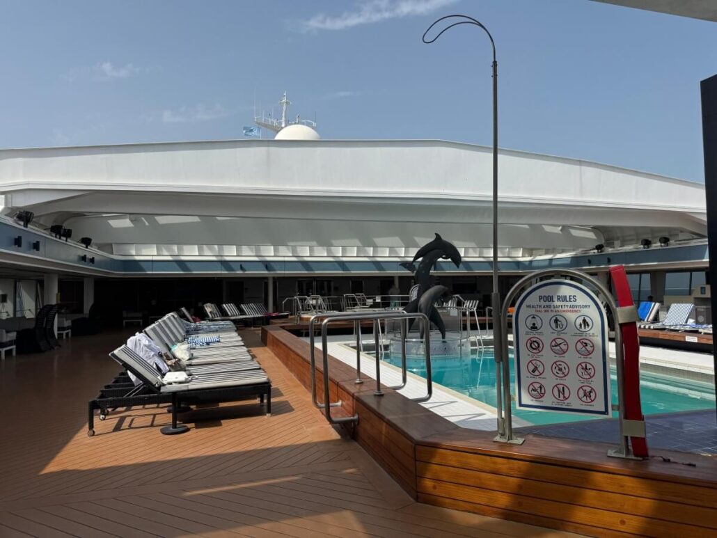 The pool deck of the Celestyal Journey is serene, offering lounge chairs and a sparkling swimming pool with a charming dolphin statue. On this spotless Embarkation Day, the sky is clear, and a sign quietly reminds everyone of the pool rules.