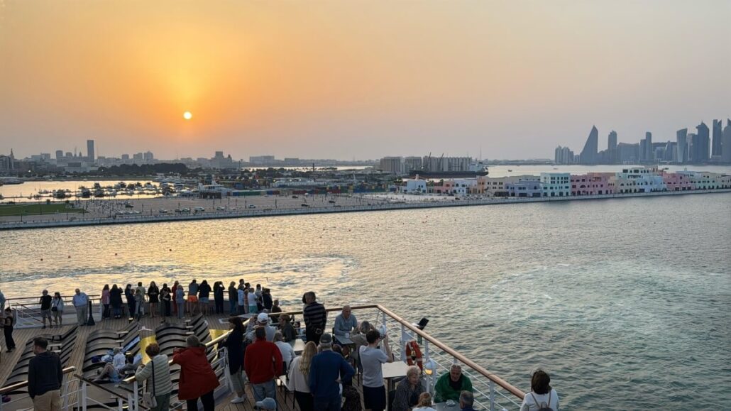 On Embarkation Day aboard the Celestyal Journey, people gather on the viewing deck to watch the sunset over Doha's skyline. The modern cityscape and sunlit waters create a stunning blend of natural beauty and urban elegance.