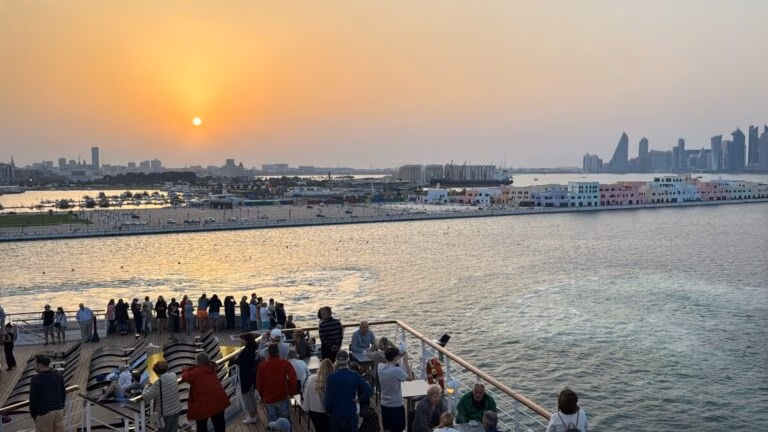 On Embarkation Day aboard the Celestyal Journey, people gather on the viewing deck to watch the sunset over Doha's skyline. The modern cityscape and sunlit waters create a stunning blend of natural beauty and urban elegance.