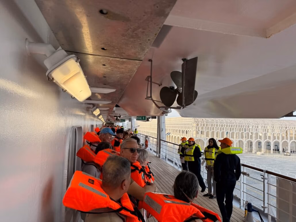 On embarkation day of the Celestyal Journey in Doha, a group wearing orange life jackets stands on the deck beneath a metal overhang. Some gaze outward at the view, while others chat. Nearby, crew members in yellow vests and helmets ensure everything runs smoothly.