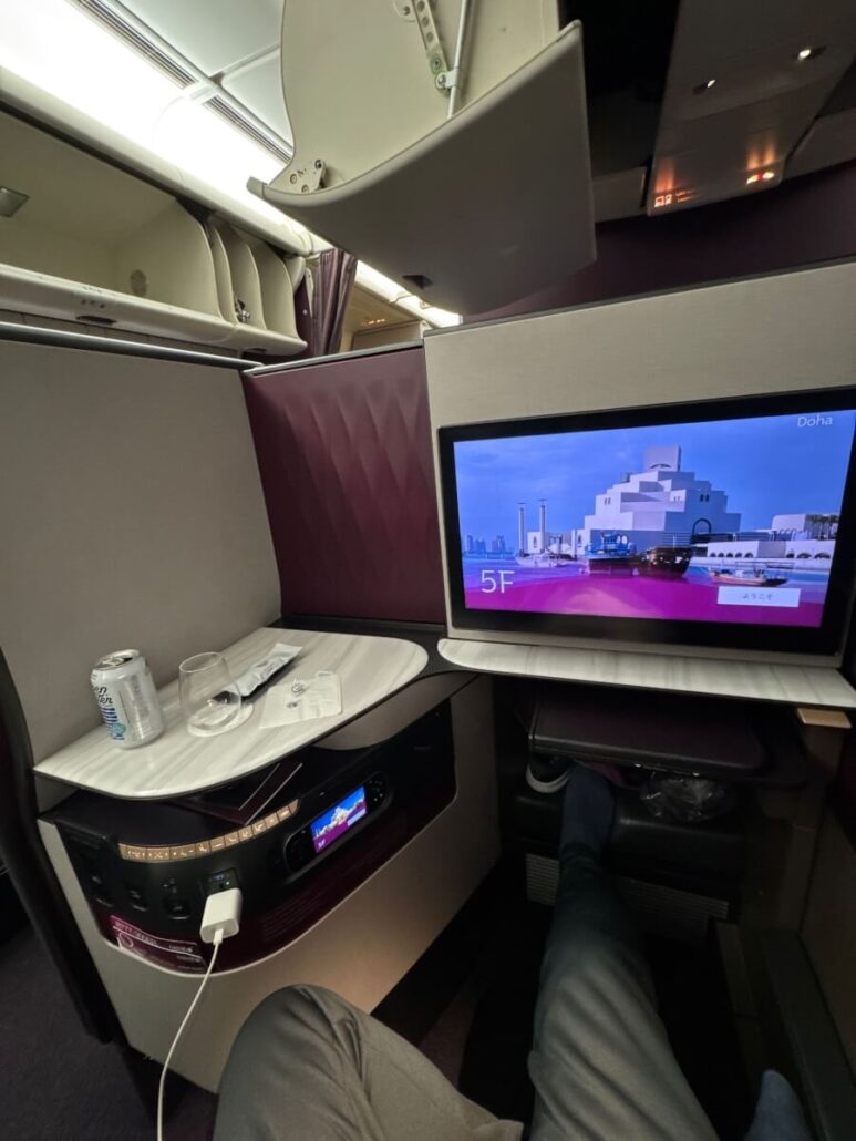 An airline passenger's view of a business class seat with a personal screen displaying a Doha cityscape. A tray table holds a glass of water and a soda can. An open overhead compartment is above, and the passenger's legs are stretched out, ready for Celestyal Journey's Embarkation Day.