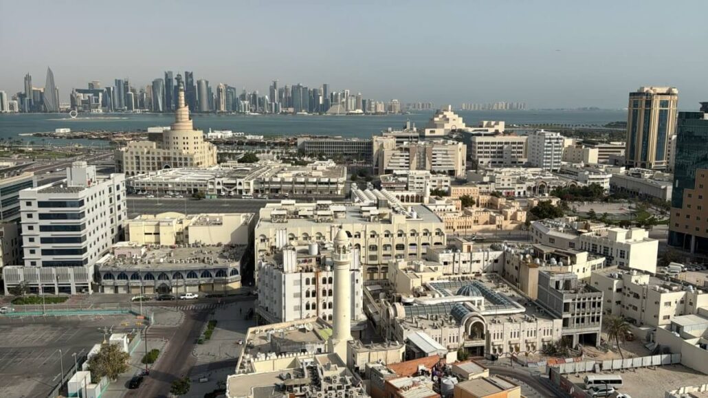 Aerial view of Doha's cityscape reveals a blend of modern and traditional buildings. In the foreground, a distinctive spiral minaret stands out, while the skyline features towering skyscrapers lining the waterfront. The sky is clear, setting a perfect scene for Embarkation Day on the Celestyal Journey.