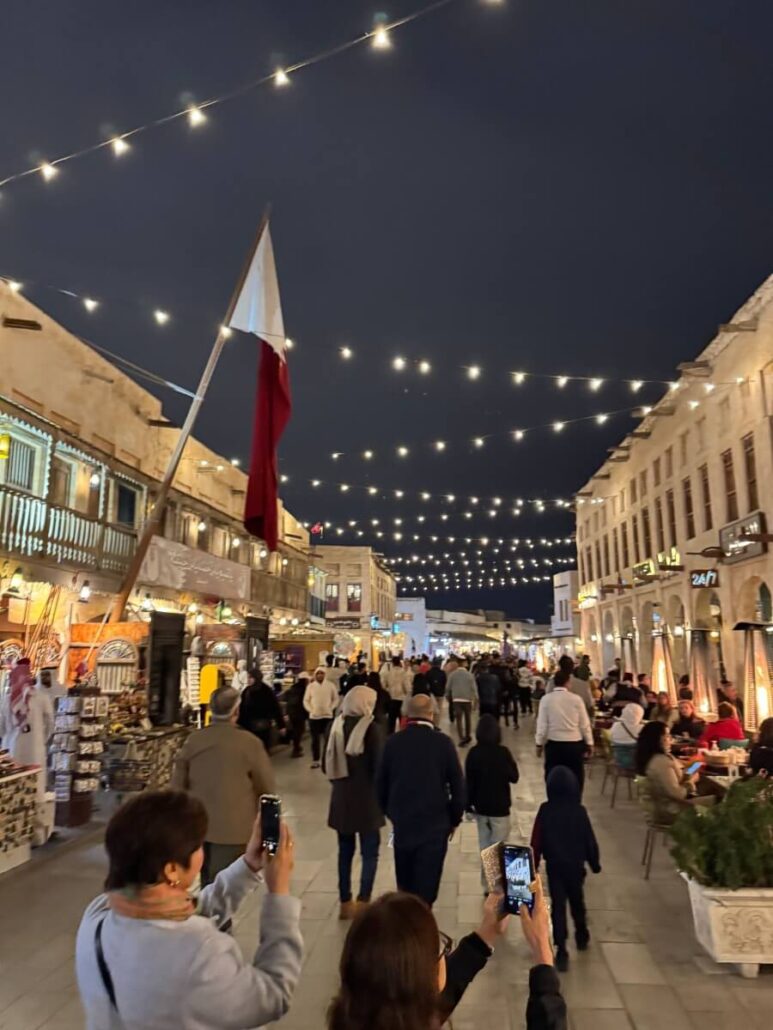 A bustling market street at night teems with people walking and dining. String lights, reminiscent of those on Celestyal Cruises exploring the Arabian Gulf, illuminate the scene. A large flag and various shop signs line the cobblestone path as individuals capture memories of a magical evening.