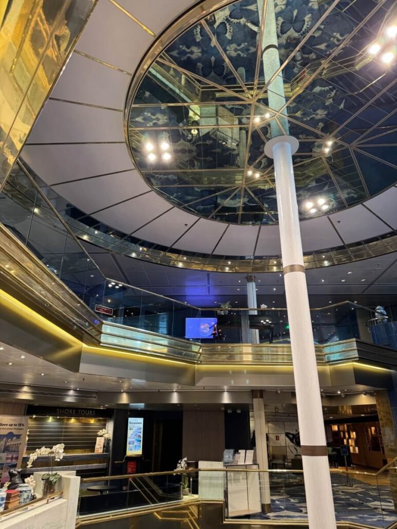 The interior of the multi-level cruise ship atrium on the Celestyal Journey dazzles with gold accents and a large, central pillar. On Embarkation Day, guests marvel at the ceiling's decorative design with glass elements. Digital screens and inviting seating areas complete this opulent space.