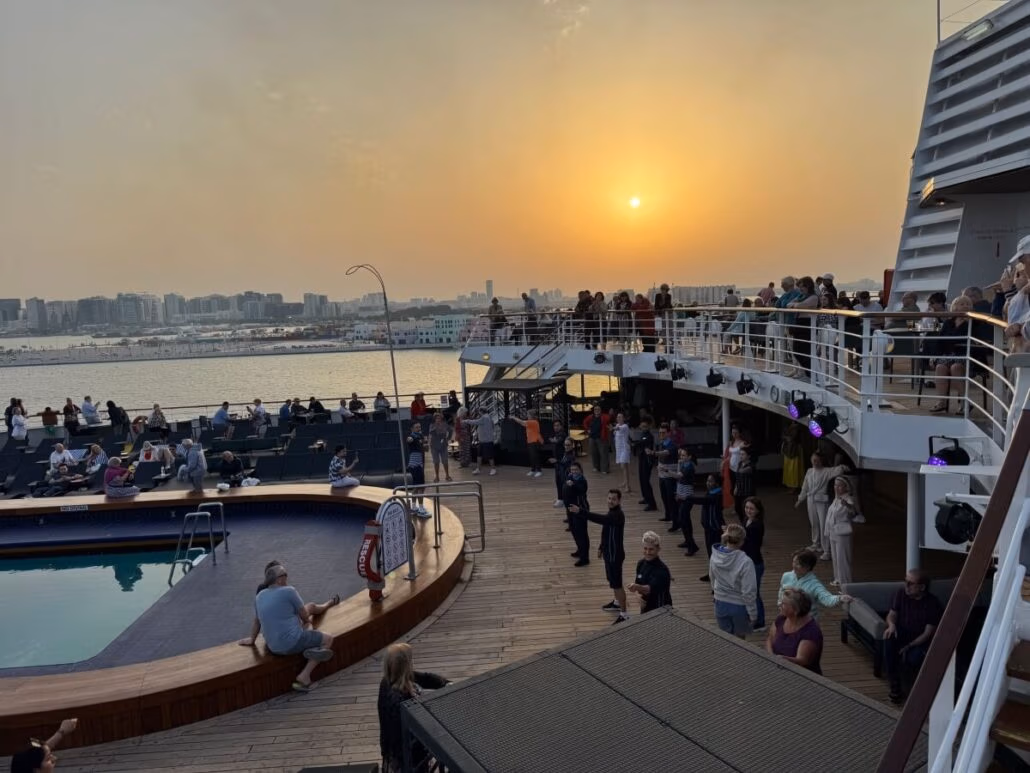On Embarkation Day, people gather on the decks of the Celestyal Journey cruise ship during sunset. The scene includes a pool, lifebuoy, and several levels of decks with railings. The skyline of Doha is visible in the background under a hazy sky.