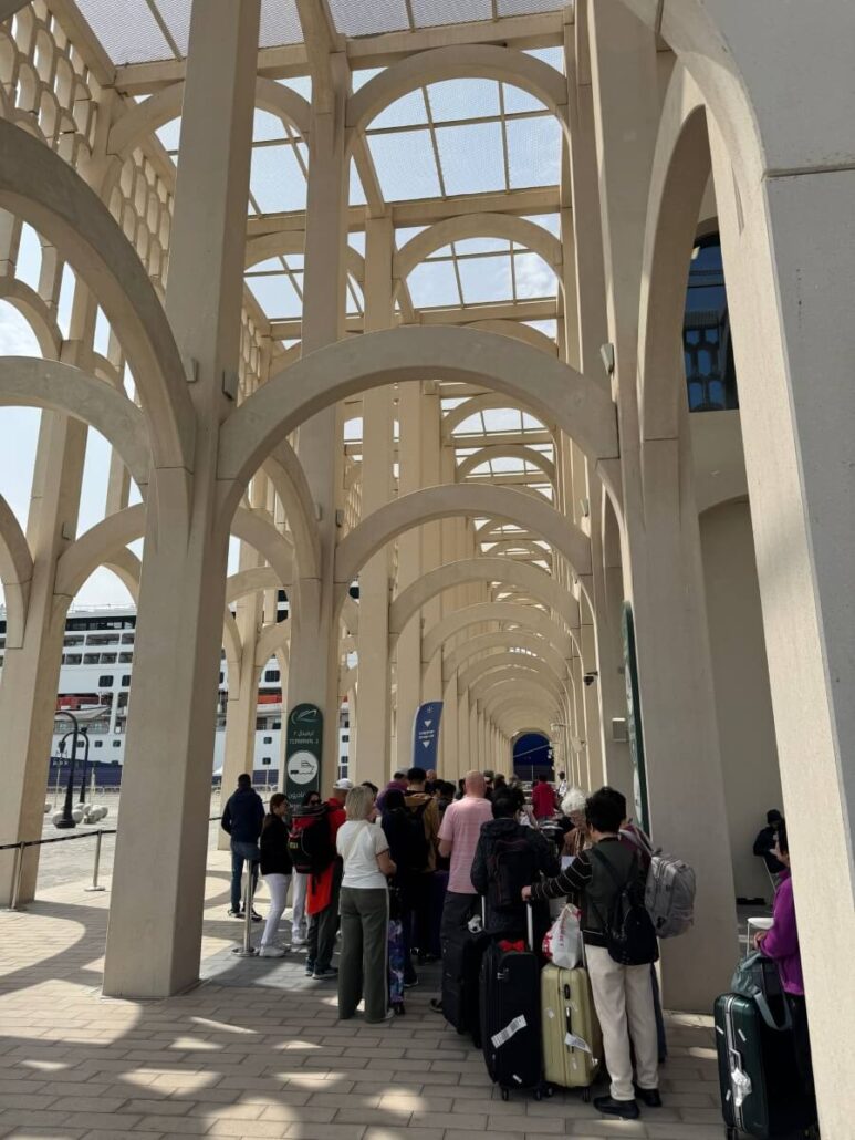 On Embarkation Day, a group of people with luggage waits in line under grand, decorative arches. The shadows dance on the stone walkway as they anticipate their Celestyal Journey. The sky is clear, and buildings punctuate the horizon in the background.
