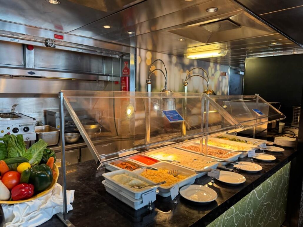 On Embarkation Day of the Celestyal Journey, a buffet setup awaits, showcasing trays of pasta and vegetables under a sleek sneeze guard. Plates are neatly stacked at the counter's end, while the stainless steel kitchen gleams in the background with fresh produce bowls adding vibrant flair.
