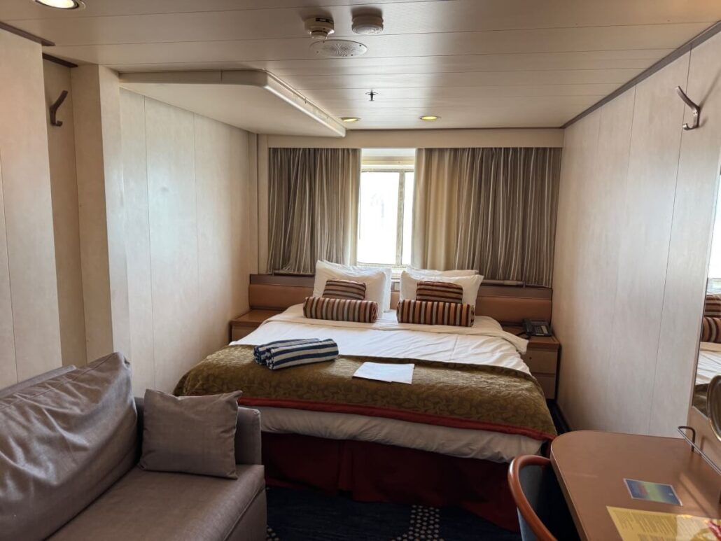 A small Celestyal Journey cruise ship cabin features a bed with striped pillows and white bedding, with a folded towel at the foot. A sofa sits on the left, a desk with a chair on the right, and a window with curtains graces the backdrop—perfect for Embarkation Day.