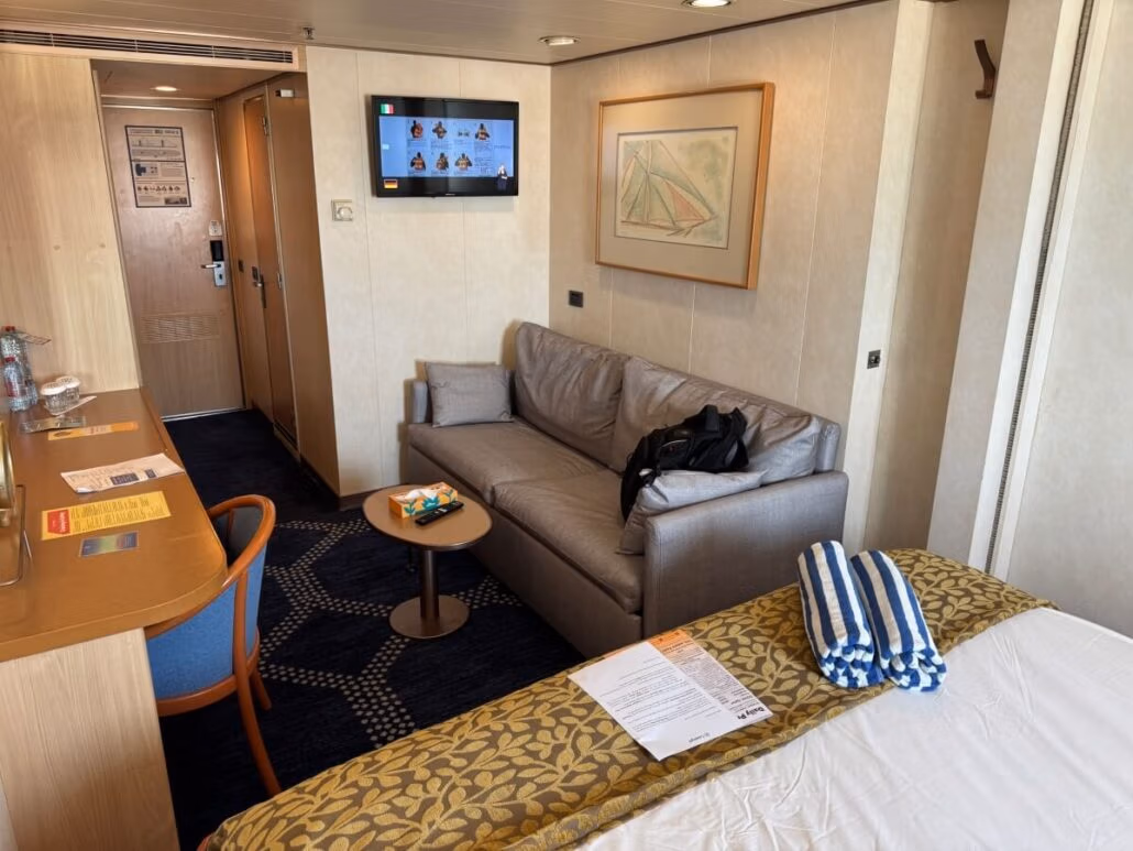 The interior of the cozy room sets the stage for embarkation day on your Celestyal Journey. A bed with striped towels, a sofa with a backpack beneath vibrant artwork, and a wall-mounted TV create a welcoming space. To the left, a table with chairs awaits. Doors lead to both hallway and bathroom adventures beyond.