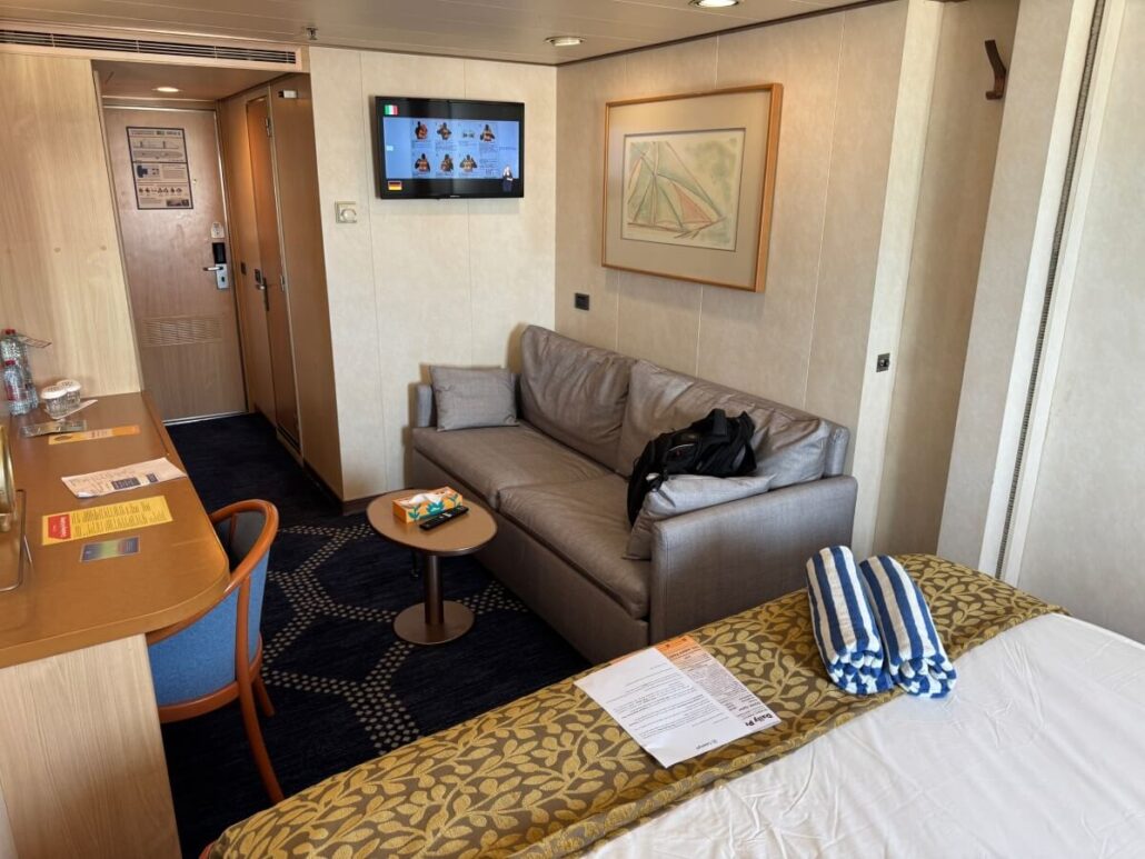 The interior of the cozy room sets the stage for embarkation day on your Celestyal Journey. A bed with striped towels, a sofa with a backpack beneath vibrant artwork, and a wall-mounted TV create a welcoming space. To the left, a table with chairs awaits. Doors lead to both hallway and bathroom adventures beyond.