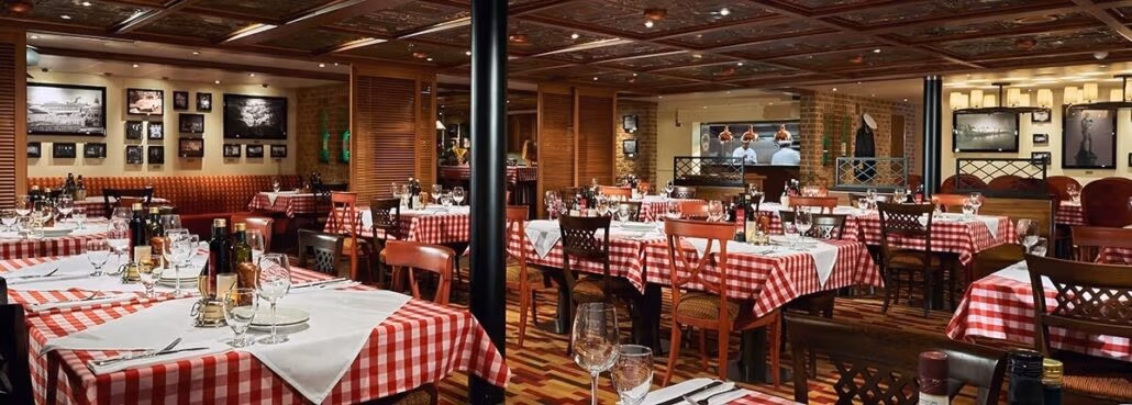 A restaurant interior featuring tables with red and white checkered tablecloths, reminiscent of carnival cruise menus, neatly set with glasses and utensils. The walls are adorned with framed pictures, and there is a visible kitchen in the back. Warm lighting creates a cozy atmosphere.