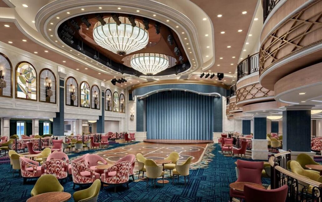 An elegant theater interior aboard the cruise showcases a central stage framed by blue curtains. The ceiling dazzles with grand chandeliers, while guests enjoy entertainment from patterned armchairs and tables on a plush blue carpeted floor. This venue offers an exquisite guide to relaxation at sea.