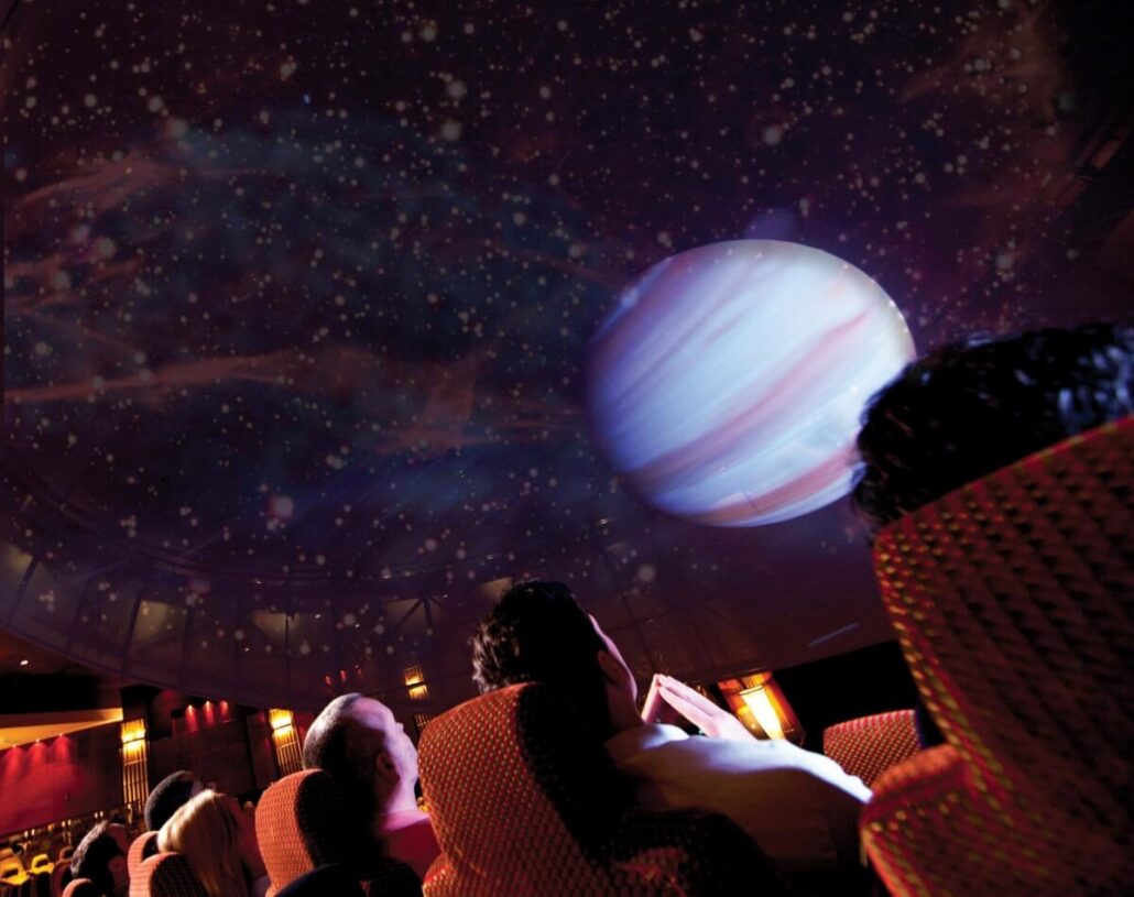 People are seated in a planetarium with red cushioned chairs, gazing up at a cosmic display projected on the dome ceiling. The dimly lit setting enhances the celestial spectacle—an unforgettable highlight featured in our exclusive Cruise Entertainment Guide.