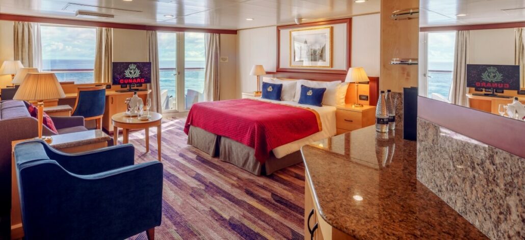 Luxurious Cunard cruise ship cabin with a large bed, red bedspread, and decorative pillows. This Queen Elizabeth room features a sitting area with armchairs, a wall-mounted TV, and a balcony overlooking the ocean. Warm lighting and modern decor enhance the cozy atmosphere for its Alaska debut.