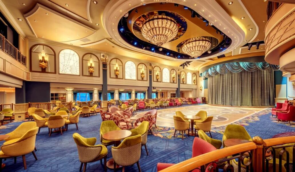 A luxurious theater lounge with a circular stage and large curtain, reminiscent of a grand debut fit for Queen Elizabeth. It's surrounded by colorful, plush chairs and tables, while ornate chandeliers and decorative lighting create an elegantly refurbished ambiance.