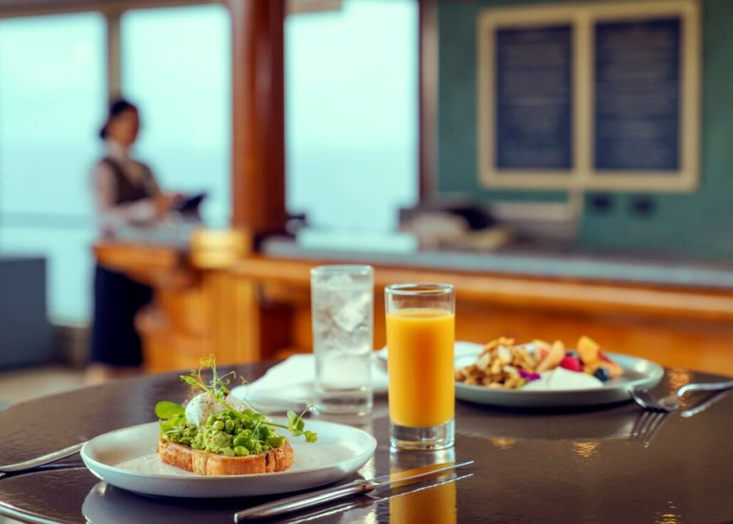 A table set for a meal with avocado toast topped with a poached egg and greens, showcasing a fresh refurbishment of classic brunch favorites. A glass of orange juice accompanies a salad of chopped fruits and nuts. A glass of water sits nearby while someone hovers by the chalkboard menu.