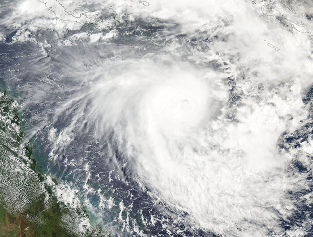 A satellite image captures a tropical cyclone's swirling cloud formations over the ocean. Dense white clouds form a circular pattern, indicating its formidable presence. Thinner clouds surround it, while a slice of land is visible at the bottom left—a scene prompting itinerary changes and cruise cancellations.