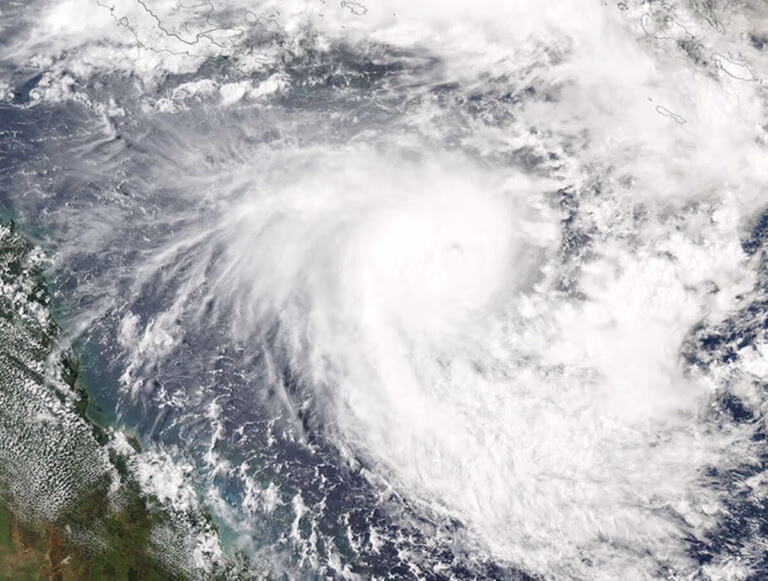 A satellite image captures a tropical cyclone's swirling cloud formations over the ocean. Dense white clouds form a circular pattern, indicating its formidable presence. Thinner clouds surround it, while a slice of land is visible at the bottom left—a scene prompting itinerary changes and cruise cancellations.