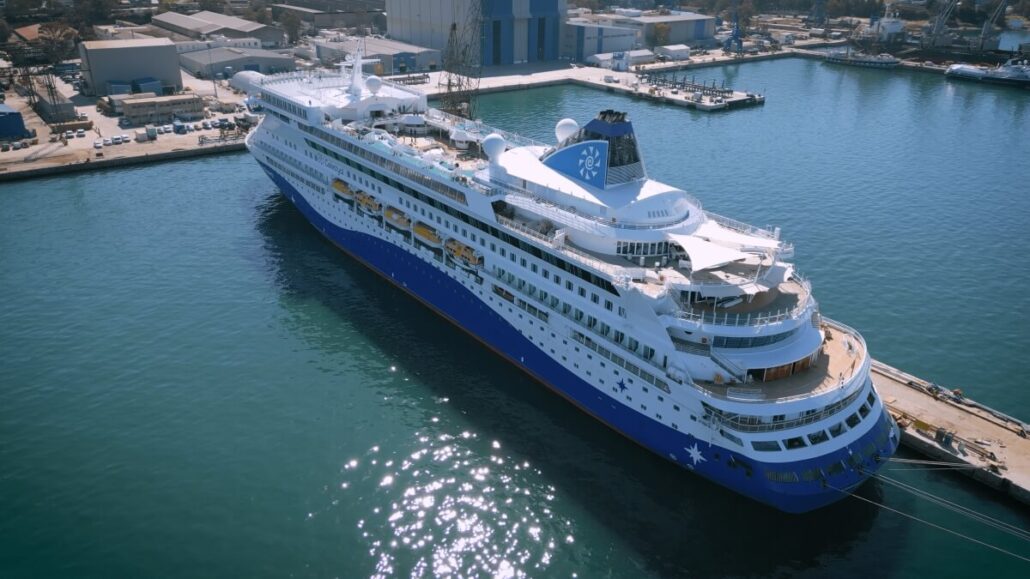 The Celestyal Discovery, with its blue and white hull and multiple decks, has returned to service. Featuring premium accommodations, the top deck boasts lifeboats and a pool area. Industrial buildings dot the background, all reflected in the calm waters of the port.