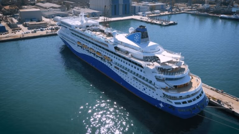 The Celestyal Discovery, with its blue and white hull and multiple decks, has returned to service. Featuring premium accommodations, the top deck boasts lifeboats and a pool area. Industrial buildings dot the background, all reflected in the calm waters of the port.