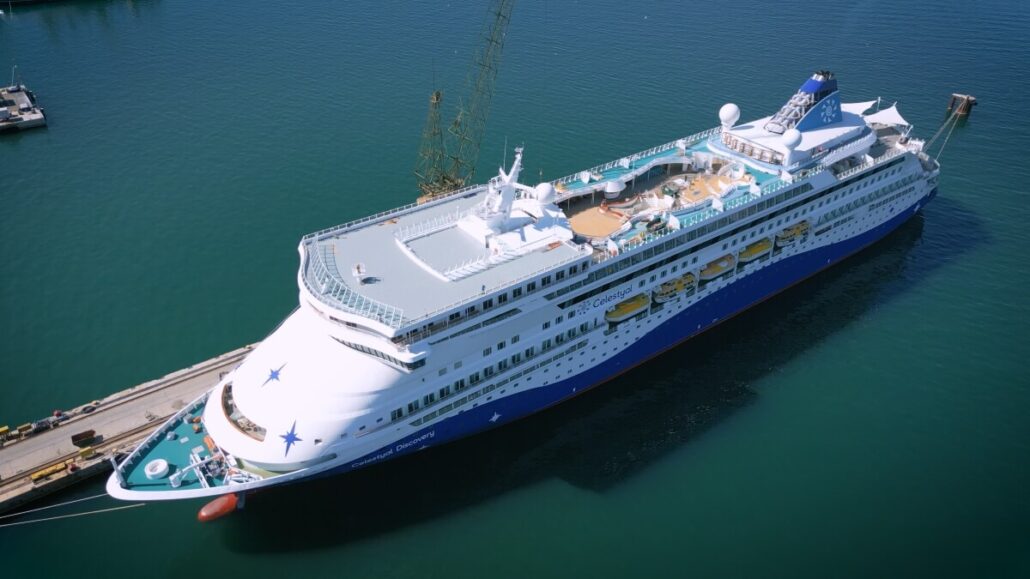Aerial view of the Celestyal Discovery cruise ship returning to service, docked at a port. The vessel boasts a sleek white and blue exterior with premium accommodations, including a sun deck and swimming pool area on the upper deck. The surrounding water is calm, and the sky is clear.