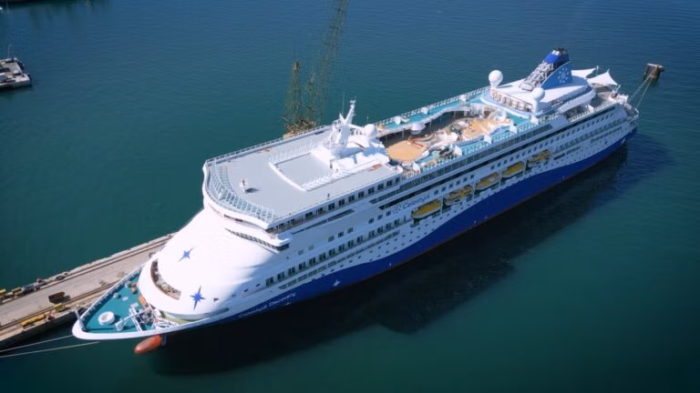 Aerial view of the Celestyal Discovery cruise ship returning to service, docked at a port. The vessel boasts a sleek white and blue exterior with premium accommodations, including a sun deck and swimming pool area on the upper deck. The surrounding water is calm, and the sky is clear.