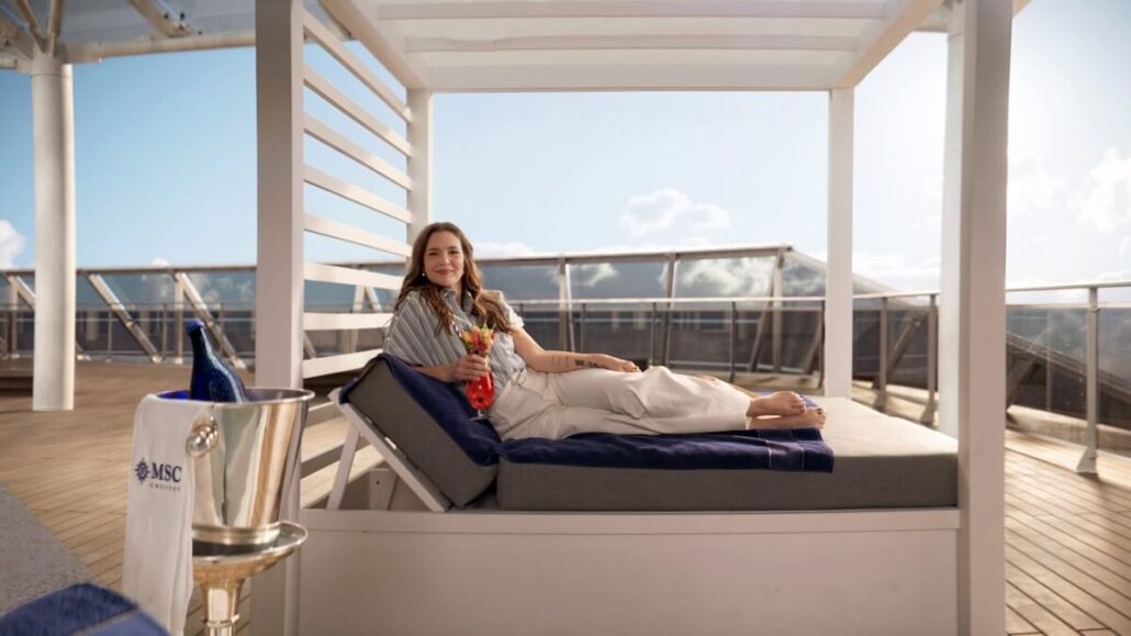 A woman is lounging on a cushioned daybed on the sunlit deck of the MSC World America. She's holding a colorful drink and smiling, with a towel beside her. A bottle in an ice bucket sits nearby, against the backdrop of a clear sky and railing. She looks like Drew Barrymore’s godmother basking in bliss.