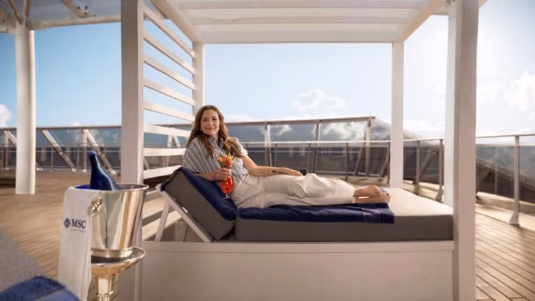 A woman is lounging on a cushioned daybed on the sunlit deck of the MSC World America. She's holding a colorful drink and smiling, with a towel beside her. A bottle in an ice bucket sits nearby, against the backdrop of a clear sky and railing. She looks like Drew Barrymore’s godmother basking in bliss.