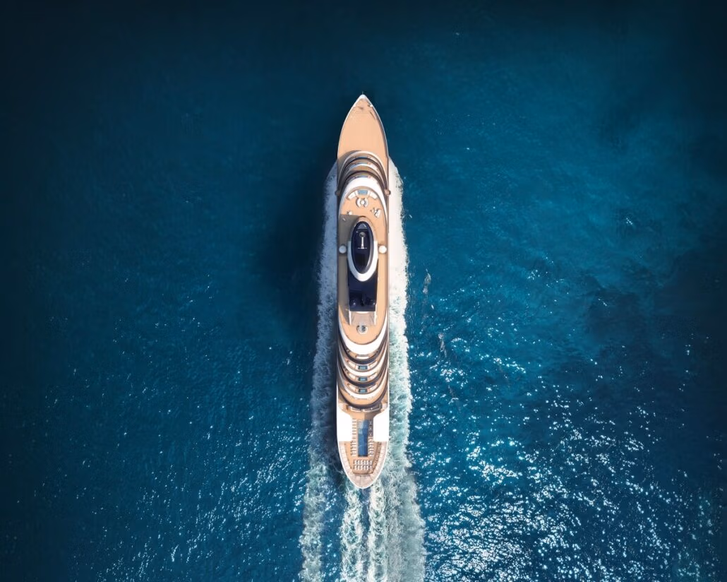 Aerial view of a Four Seasons Yacht cruising through clear blue ocean water, expertly helmed by Captain Kate McCue. The yacht creates a white wake as it moves, indicating speed. The sun reflects off the water, creating a bright, serene atmosphere.
