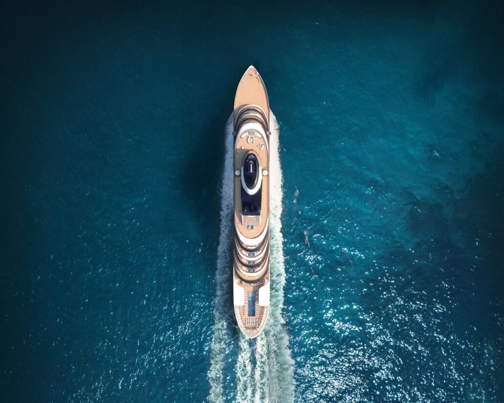 Aerial view of a Four Seasons Yacht cruising through clear blue ocean water, expertly helmed by Captain Kate McCue. The yacht creates a white wake as it moves, indicating speed. The sun reflects off the water, creating a bright, serene atmosphere.