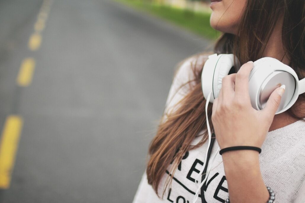 A person with long brown hair stands on a road, captured in a pensive mood. Wearing a white shirt, they hold large white headphones around their neck, ready to cruise into their favorite tunes. The empty road and yellow lines echo freedom; only part of the face is shown.