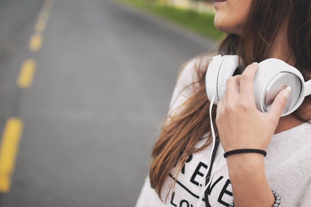 A person with long brown hair stands on a road, captured in a pensive mood. Wearing a white shirt, they hold large white headphones around their neck, ready to cruise into their favorite tunes. The empty road and yellow lines echo freedom; only part of the face is shown.