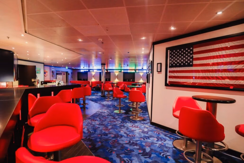 The vibrant modern lounge aboard the Carnival Spirit features bright red chairs and round tables set on a patterned carpet. Enhanced service meets guests under a ceiling lined with lights, alongside a proudly displayed American flag. To the left, a sleek bar area invites relaxation.