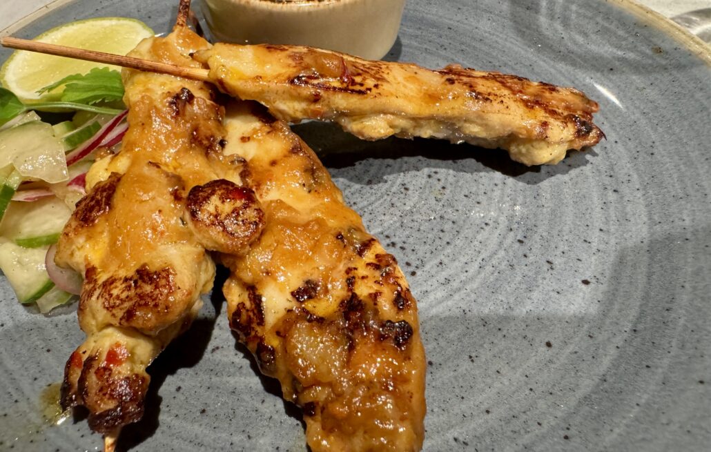Two grilled satay chicken skewers with a golden glaze are served on a gray plate aboard Norwegian Cruise Line's latest ship. They are accompanied by a side salad with lettuce, cucumbers, and radishes, plus a lime wedge. A small dish of dipping sauce is partially visible in the background.