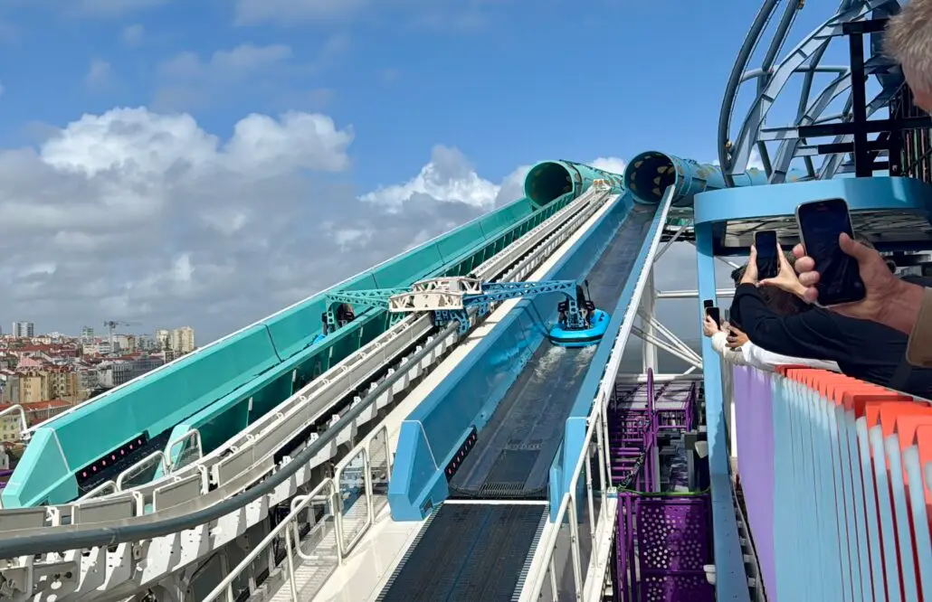 The Norwegian Aqua slidecoaster with a raft ascends toward a tunnel entrance on the Norwegian Aqua, NCL's latest ship. The slide is surrounded by railings and a viewing platform where people capture photos. In the background, the cityscape stands against a partly cloudy sky.