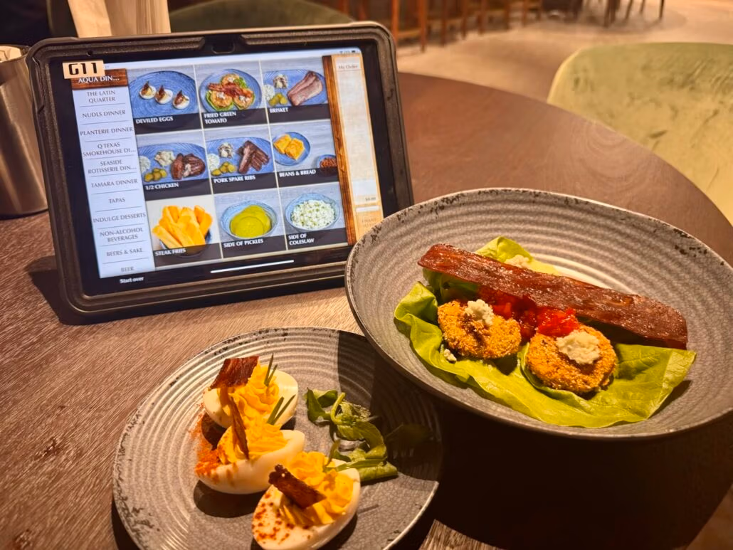 A plate with deviled eggs topped with bacon, alongside fried green tomatoes with a sauce, rests on a bed of greens. Next to it, an iPad shows the menu from NCL's latest ship featuring Norwegian Aqua cuisine with enticing food images and descriptions.