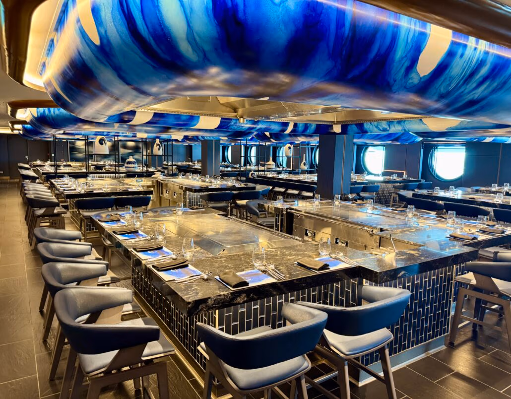 Discover a modern restaurant interior inspired by Norwegian Aqua, with a central square dining area featuring a granite countertop and built-in grills. Blue cushioned chairs provide comfort while overhead, blue and white lighting structures offer a vibrant touch reminiscent of NCL's latest ship design.