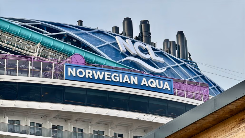 The image showcases the upper section of NCL's latest ship, Norwegian Aqua, featuring several smokestacks. A prominent sign displays "Norwegian Aqua," while another at the top features "NCL" with a wave design.