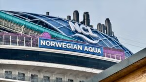 The image showcases the upper section of NCL's latest ship, Norwegian Aqua, featuring several smokestacks. A prominent sign displays "Norwegian Aqua," while another at the top features "NCL" with a wave design.