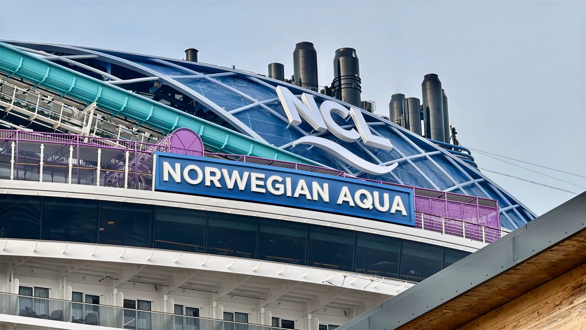 The image showcases the upper section of NCL's latest ship, Norwegian Aqua, featuring several smokestacks. A prominent sign displays "Norwegian Aqua," while another at the top features "NCL" with a wave design.