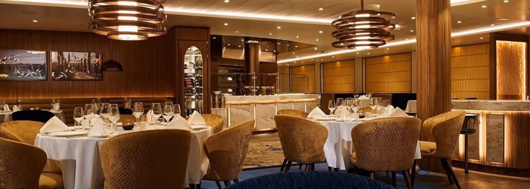 Elegant restaurant interior reminiscent of a refined ocean voyage, with warm lighting and wooden decor. Tables are set with white tablecloths, glassware, and folded napkins similar to those found on carnival cruise menus. Modern chandeliers hang from the ceiling as a bar counter is visible in the background.