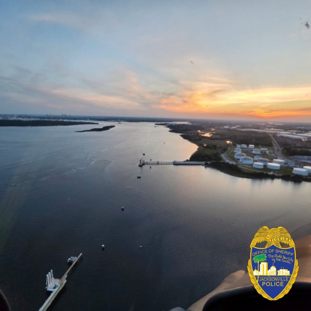 An aerial view captures a wide river lined with docks and industrial buildings at sunset, where the sky melds orange, pink, and blue. A Carnival Cruise Ship is docked nearby. In the bottom right corner, the Jacksonville Police badge is elegantly overlaid.